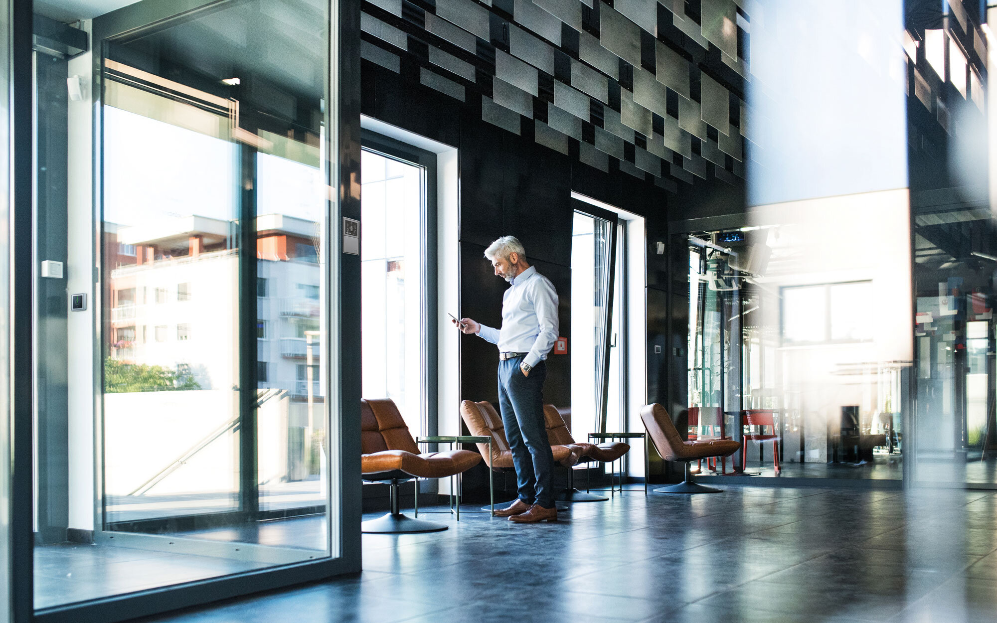 Mann mit Handy steht am Fenster eines modernen Hotelfoyers